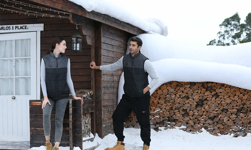 Two models wearing heated vests outside a snowy cabin